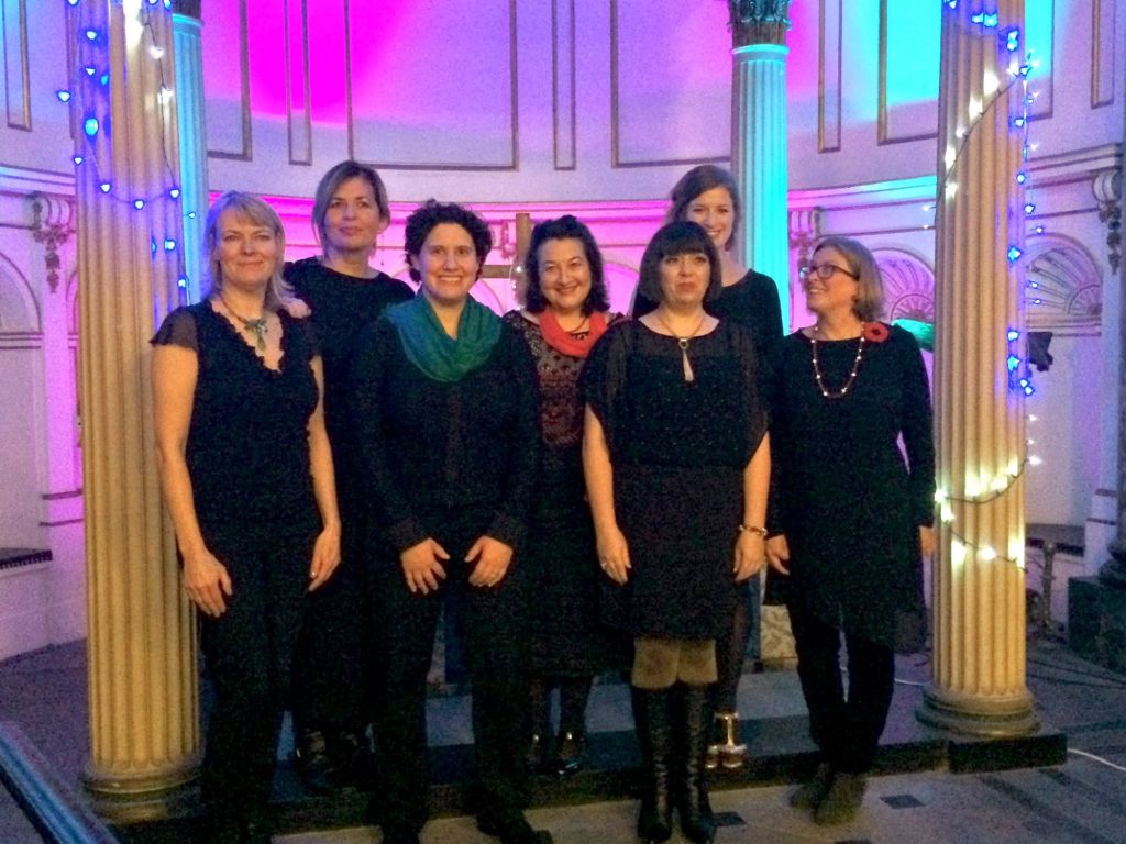 Seven singers of Rebelles Women's Choir in black, standing between two pillars on a church stage lit in pink and blue, with fairy lights around the columns.