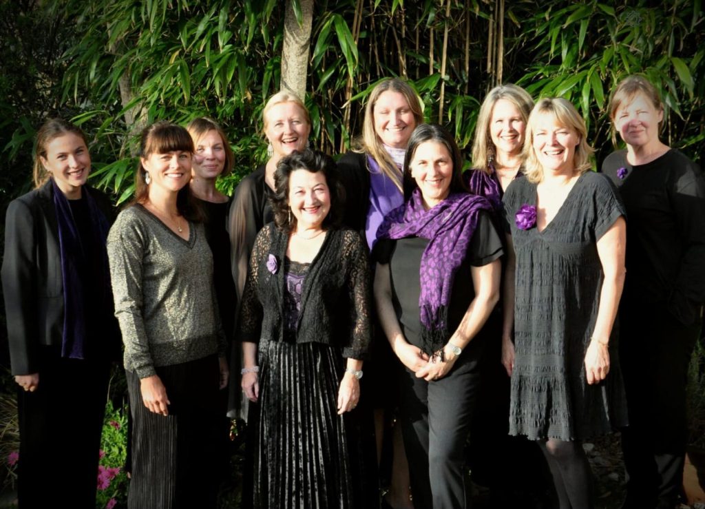 Ten singers of Rebelles Women's Choir standing together outdoors in front of bamboo, dressed in black with purple scarves and corsages.